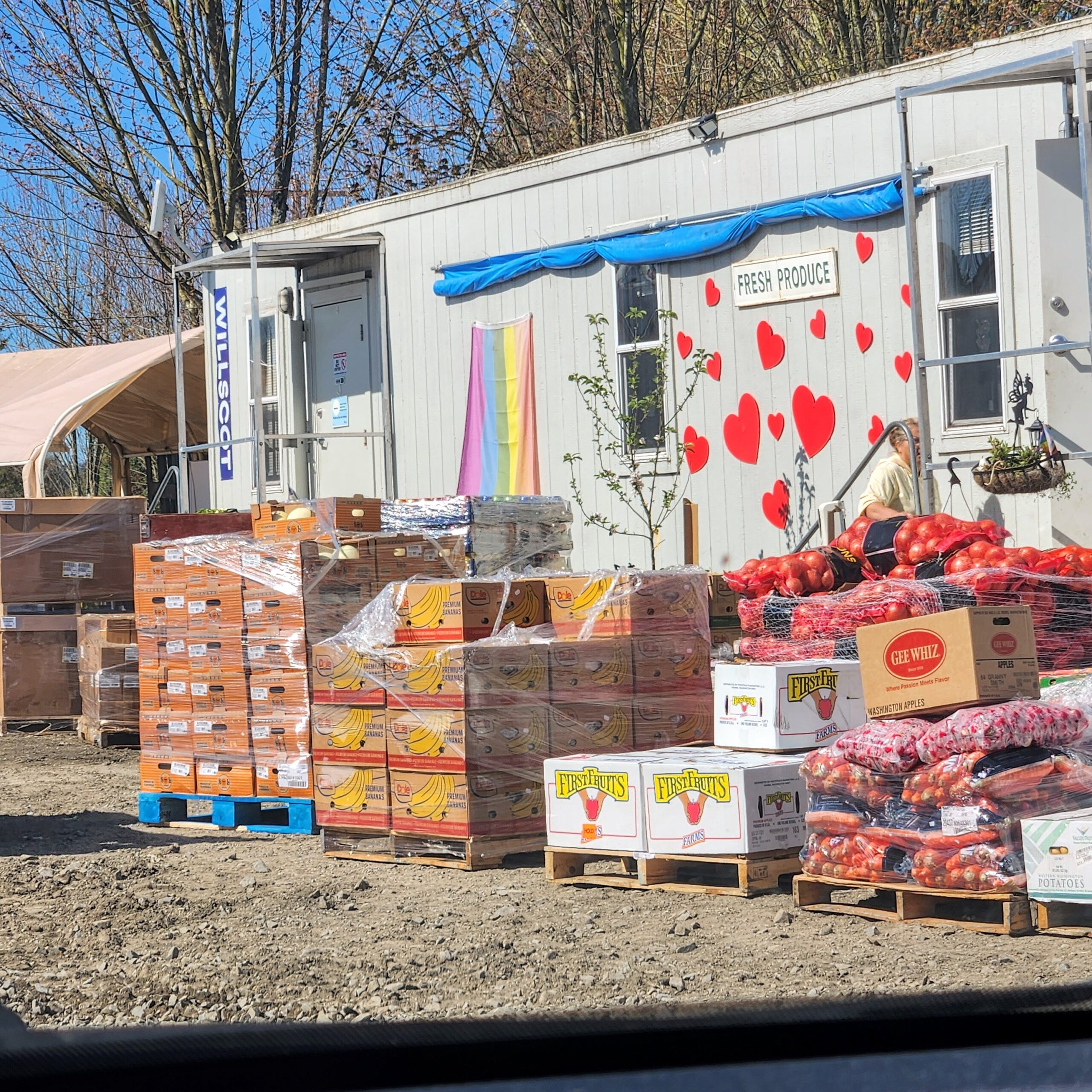 A food distribution center with several pallets of boxed fresh produce, including potatoes and onions, in front of a gray mobile office trailer decorated with a "Fresh Produce" sign and hearts. A rainbow flag hangs to the left of the door, and a person is visible behind the produce. The setting appears sunny and outdoors.