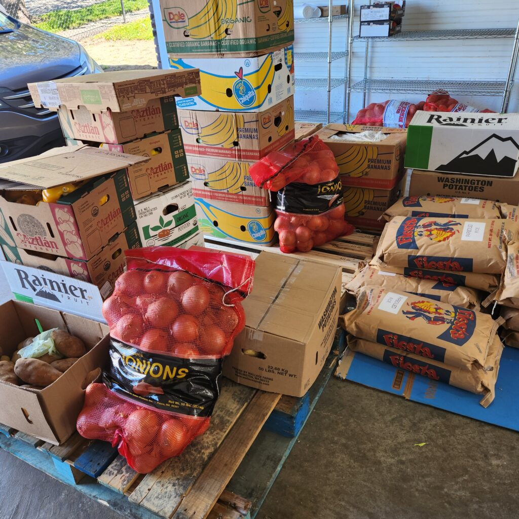 Stacks of various fresh produce boxes in a food distribution area. The boxes contain items such as organic bananas, Washington potatoes, onions, and other vegetables.